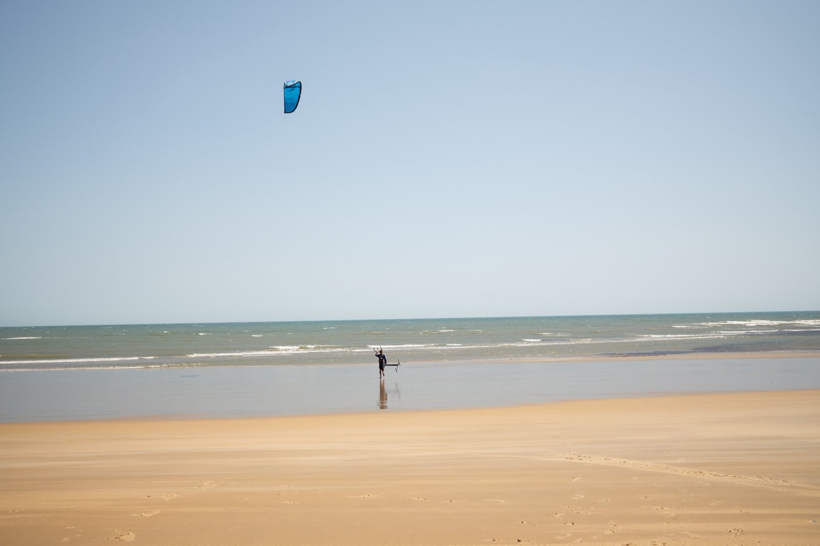A lone kitesurfer walking the wide, empty beach at Emboaca, kite flying overhead
