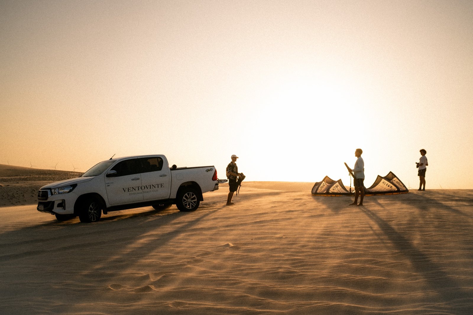 Pickup truck loaded with kite gear on golden dunes at sunset before a downwinder