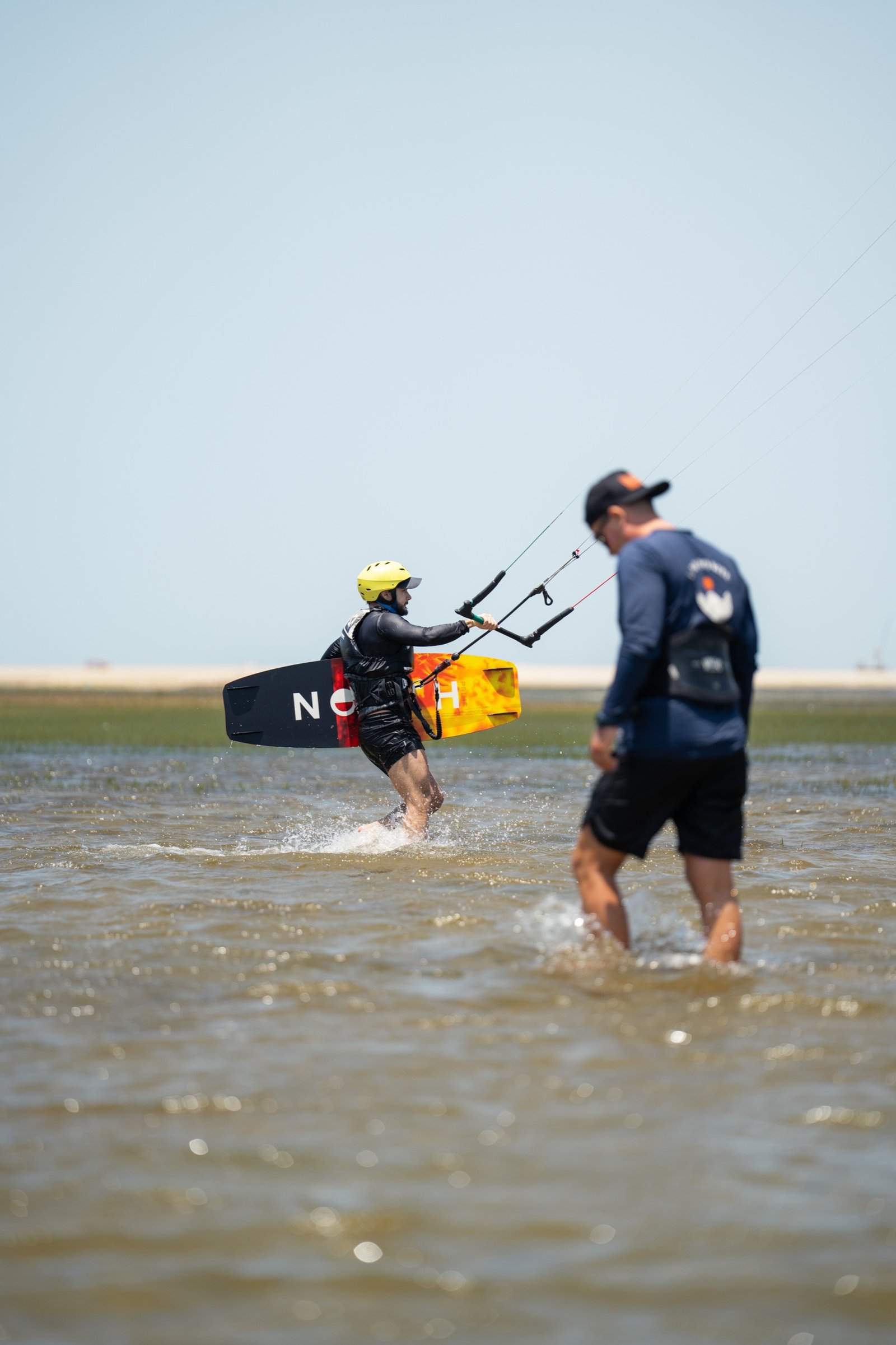 Student carrying a board through waist-deep water beside an instructor during a kitesurf lesson